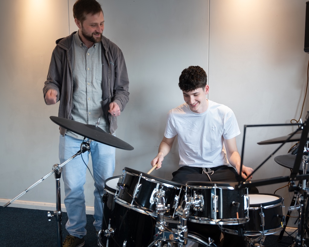 A person playing the drums, with another person stood next to them playing the air drums in an instructor kind of way. They look happy.