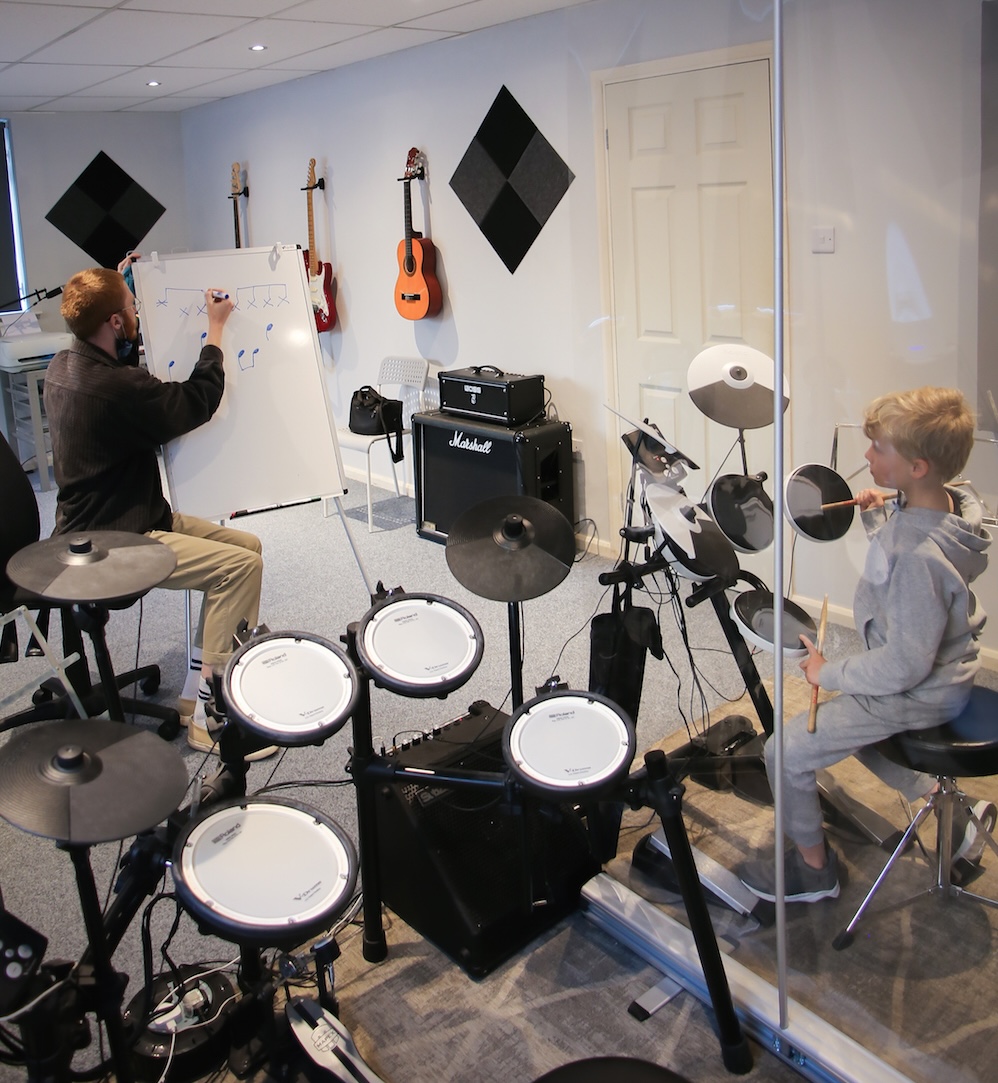 A child sat at an electric drum kit while observing a teacher who is drawing on a whiteboard. They are in a music room